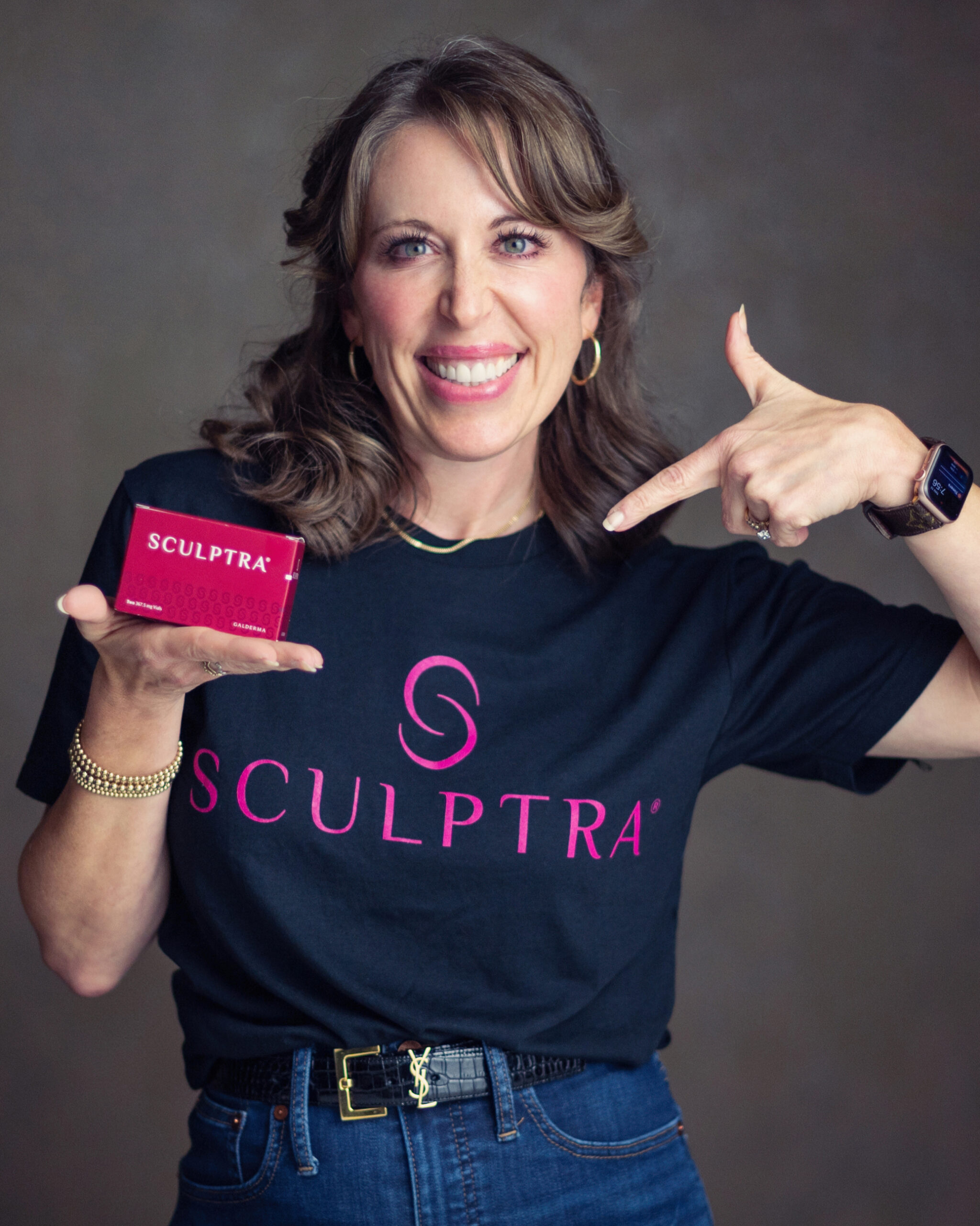 SMALKOSKI Party-190 crop Smiling woman in Racine headshot holding a pink SCULPTRA box, wearing navy shirt and gold bracelet
